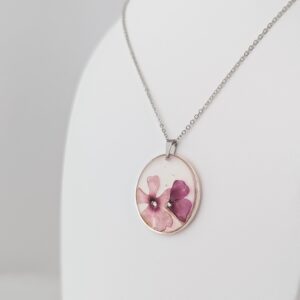PXL_20261013_184955192PORTRAIT - Blue Pendulum Jewelries Silver chain necklace with an oval pendant containing pressed pink flowers on a pale background.