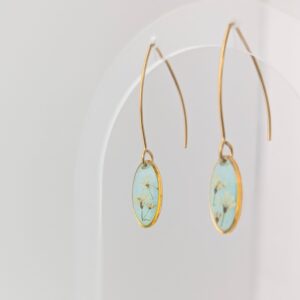 PXL_20261013_195407819PORTRAIT - Blue Pendulum Jewelries Close-up of gold hoop earrings with turquoise enamel oval pendants hanging against a pale background.