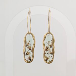 PXL_20261014_141125089PORTRAITORIGINAL - Blue Pendulum Jewelries Two gold hoop earrings with clear teardrop pendants containing dried flowers, hanging against a pale background.