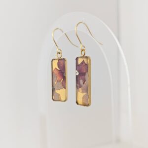 Close-up of gold hooks with rectangular pendant earrings hanging on a clear display, featuring abstract multicolor designs
