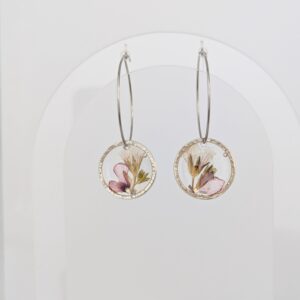 Silver hoop earrings with circular pendant charms featuring pressed flowers against a white background, hanging from a white surface.