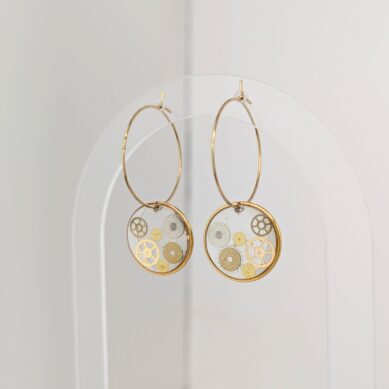 Large gold hoop earrings hanging against a pale wall, with gear-patterned inner discs visible.