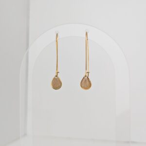 Two gold teardrop earrings hanging from clear pegs on a white curved display backdrop.